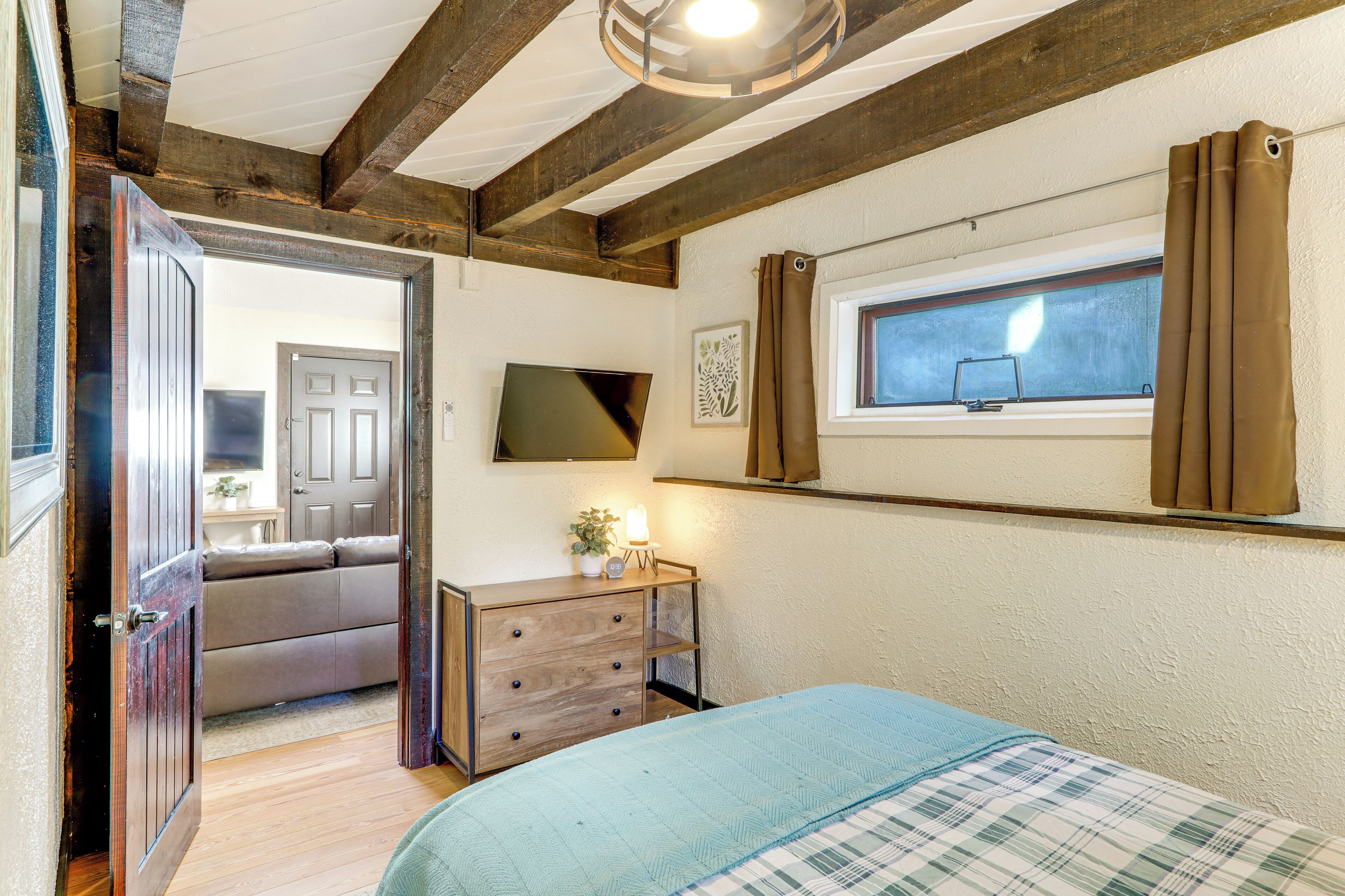 A bedroom with a bed, a bedside table, a window with curtains, a wall-mounted TV, and a door leading to another room.