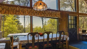 A dining area with a table set for four, wooden chairs, and a view of trees through large windows. - Hike, Bike & Golf: Peaceful Cabin in Twain Harte! (Twain Harte)