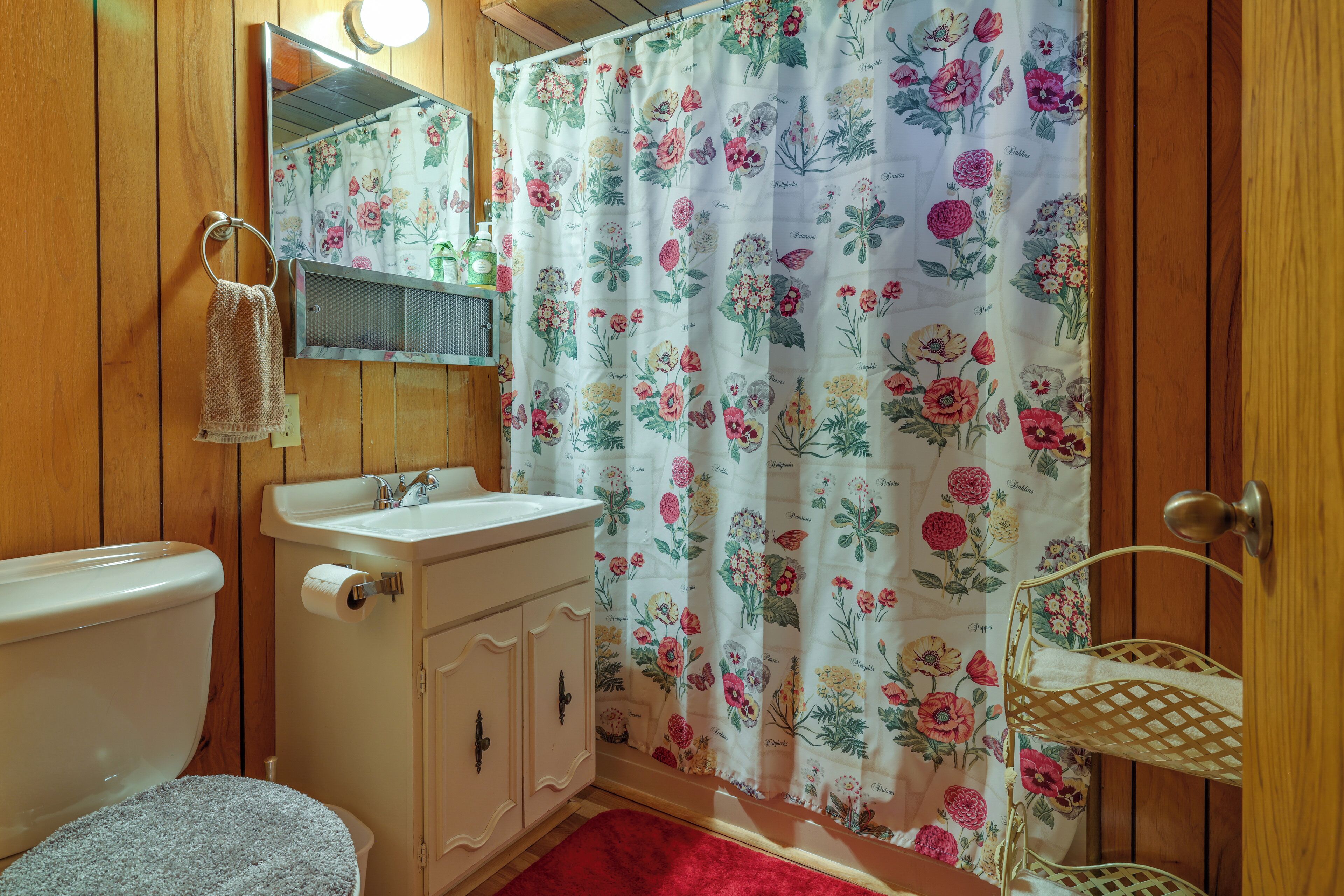 A bathroom with wooden paneling, a toilet, a sink with a mirror, a shower curtain with floral patterns, and a red bath mat.