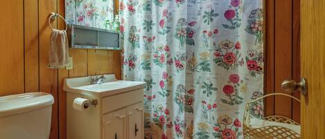 A bathroom with wooden paneling, a toilet, a sink with a mirror, a shower curtain with floral patterns, and a red bath mat.