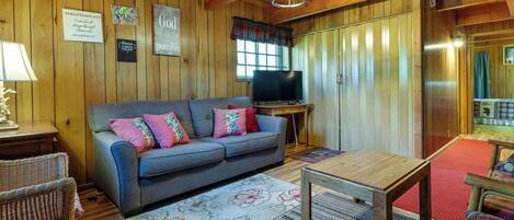 A cozy cabin interior with a wooden ceiling, a sofa with colorful pillows, a television, and a dining table.