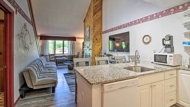 A compact kitchen with a granite countertop, white cabinets, and a dishwasher. A dining area with a sofa and chairs is visible in the background. - Resort Condo w/ Lake + Pool ~ 5 Mi to Flume Gorge! (North Woodstock)