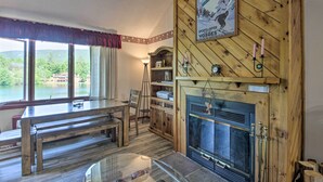 A cozy room with a fireplace, wooden walls, a dining table, and a view of a lake. - Resort Condo w/ Lake + Pool ~ 5 Mi to Flume Gorge! (North Woodstock)