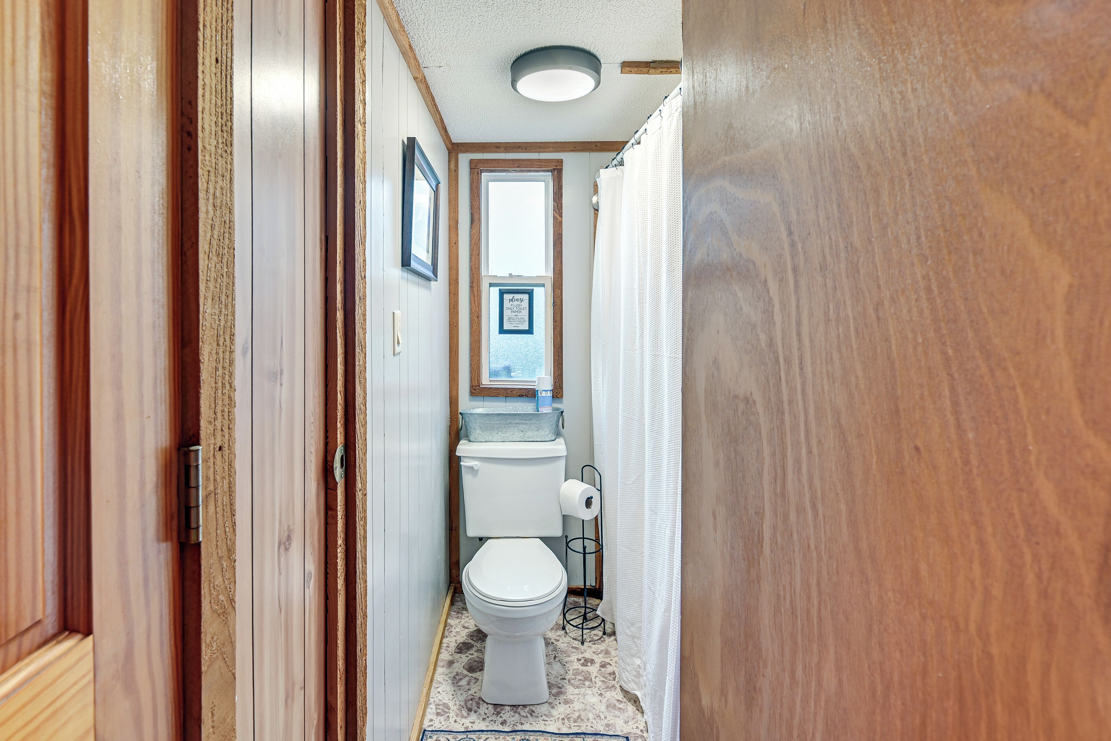 Un couloir étroit mène à une salle de bain équipée de toilettes, d’un lavabo et d’un rideau de douche.