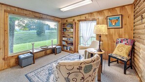 A cozy room with wooden walls, a large window, a sofa, a chair, a lamp, and a bookshelf. - Cabin on Working Highland Cattle Farm in Kentucky (Annville)