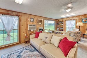 Ein gemütliches Zimmer mit Holzvertäfelung, einem beige gefärbten Sofa mit roten Kissen, einem gemusterten Teppich und einem großen Fenster mit Blick ins Freie.