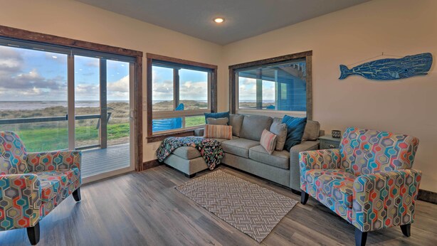 A spacious room with a large window overlooking a beach, a cozy sofa, patterned armchairs, and a decorative fish wall art piece. - Sanderling Sea Cottages, Unit 6 w/ Beach Access! (Waldport)