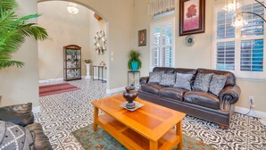 A living room with a brown leather sofa, a wooden coffee table, patterned floor, a chandelier, and a large window with shutters. - Luxe Atwater Home w/ Patio: 10 Mi to Yosemite Lake (Atwater)