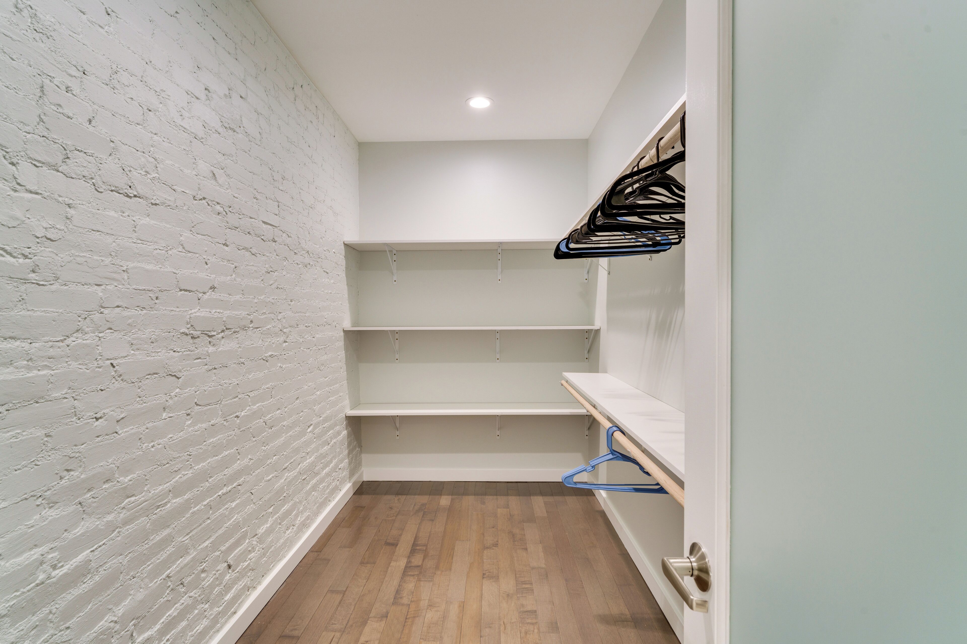 A narrow hallway with white brick walls, wooden flooring, and a white door with a metal handle. Shelves are mounted on the wall, and a blue hanger is visible.