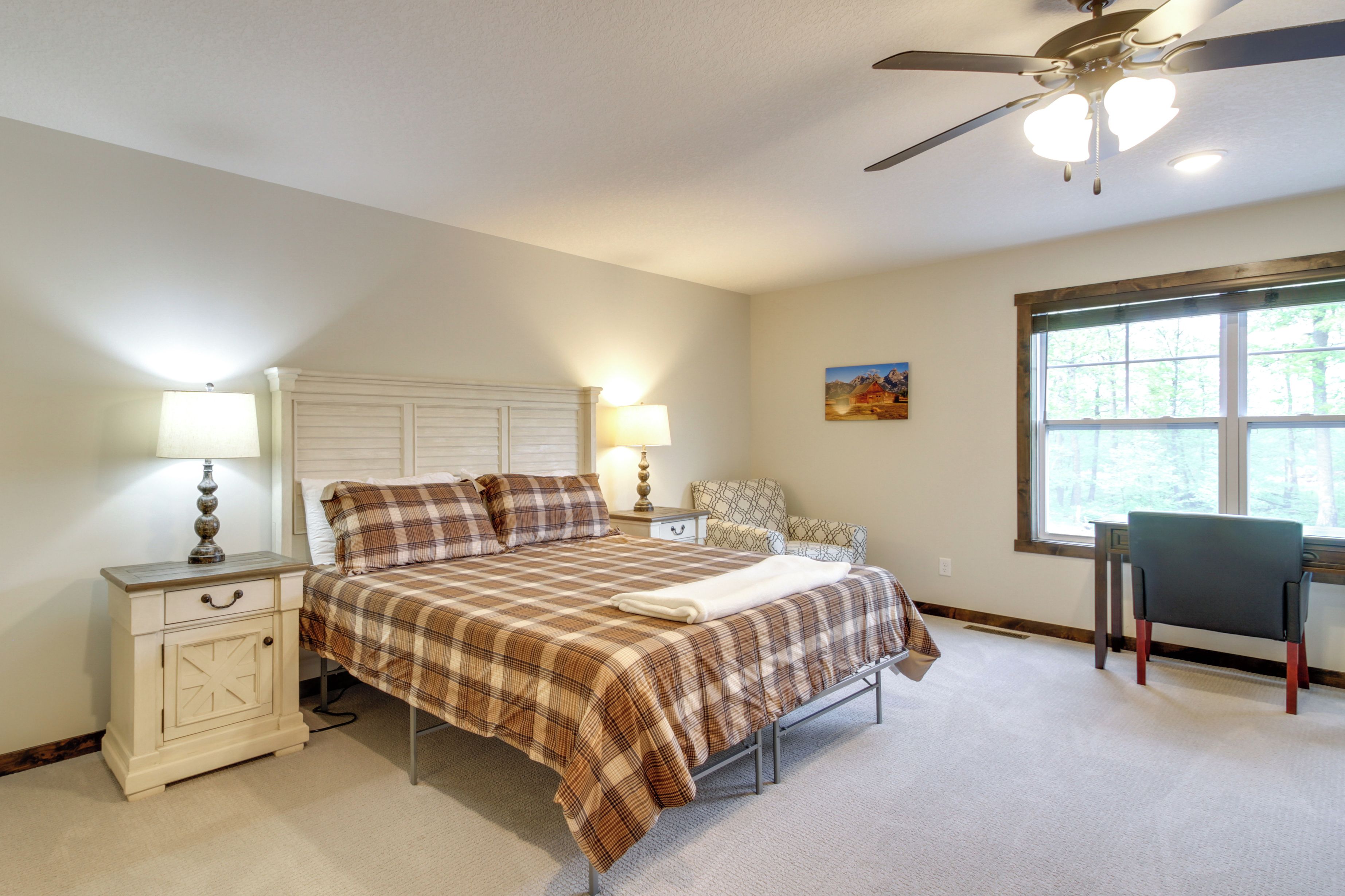 A bedroom with a bed, nightstand, lamp, chair, and a window with a view of trees.