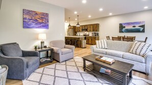 Una sala moderna con un sofá gris, un sillón gris y una mesa de centro. Se observa un área de cocina al fondo con armarios de madera y una mesa de comedor.