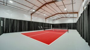Un court de tennis intérieur doté d’une surface de jeu rouge, de rideaux noirs et d’un plafond haut avec des poutres apparentes.