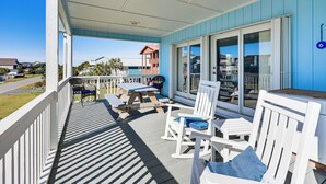 A covered outdoor deck with white furniture, a table, and a view of the ocean and neighboring houses. - For Families & Pets! Oceanside Oak Island Getaway (Oak Island)