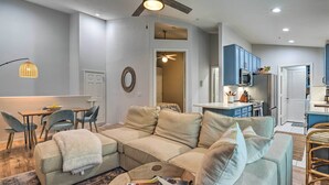 Uma sala de estar moderna com um sofá seccional, uma área de jantar com mesa e cadeiras, e uma cozinha com armários azuis e micro-ondas.