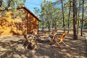 Une cabane en rondins avec un foyer et des chaises en bois disposées autour, le tout entouré d’arbres.