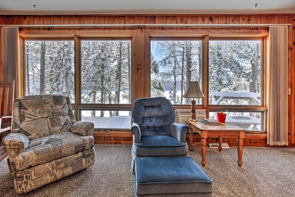 A cozy room with a large window, a blue armchair, a patterned armchair, and a wooden table with a lamp. - Serene Woodruff Cottage: Lakefront Access! (Woodruff)