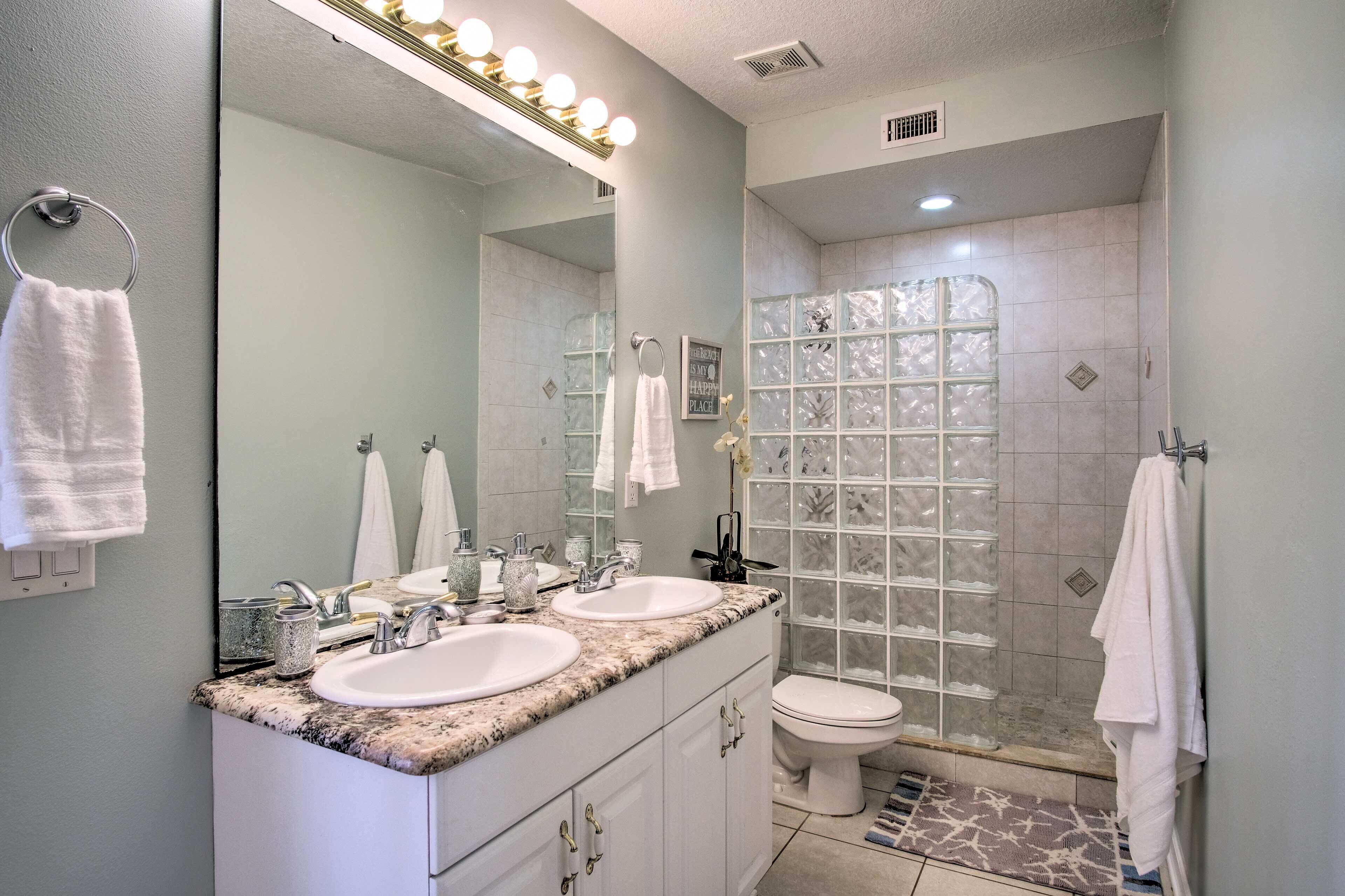 A bathroom with two sinks, a large mirror with lights, a shower with a glass door, and a toilet.