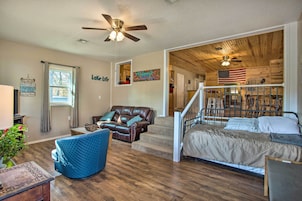 A spacious living room with a brown leather couch, a blue armchair, and a bed with a wooden headboard. A ceiling fan and a flag are visible.