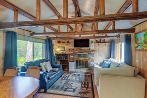 Een knus chaletinterieur met een open haard, leren banken, een eettafel en een groot raam met uitzicht op groen.