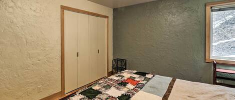 Quarto com cama, colcha, guarda-roupa e janela com vista para árvores.