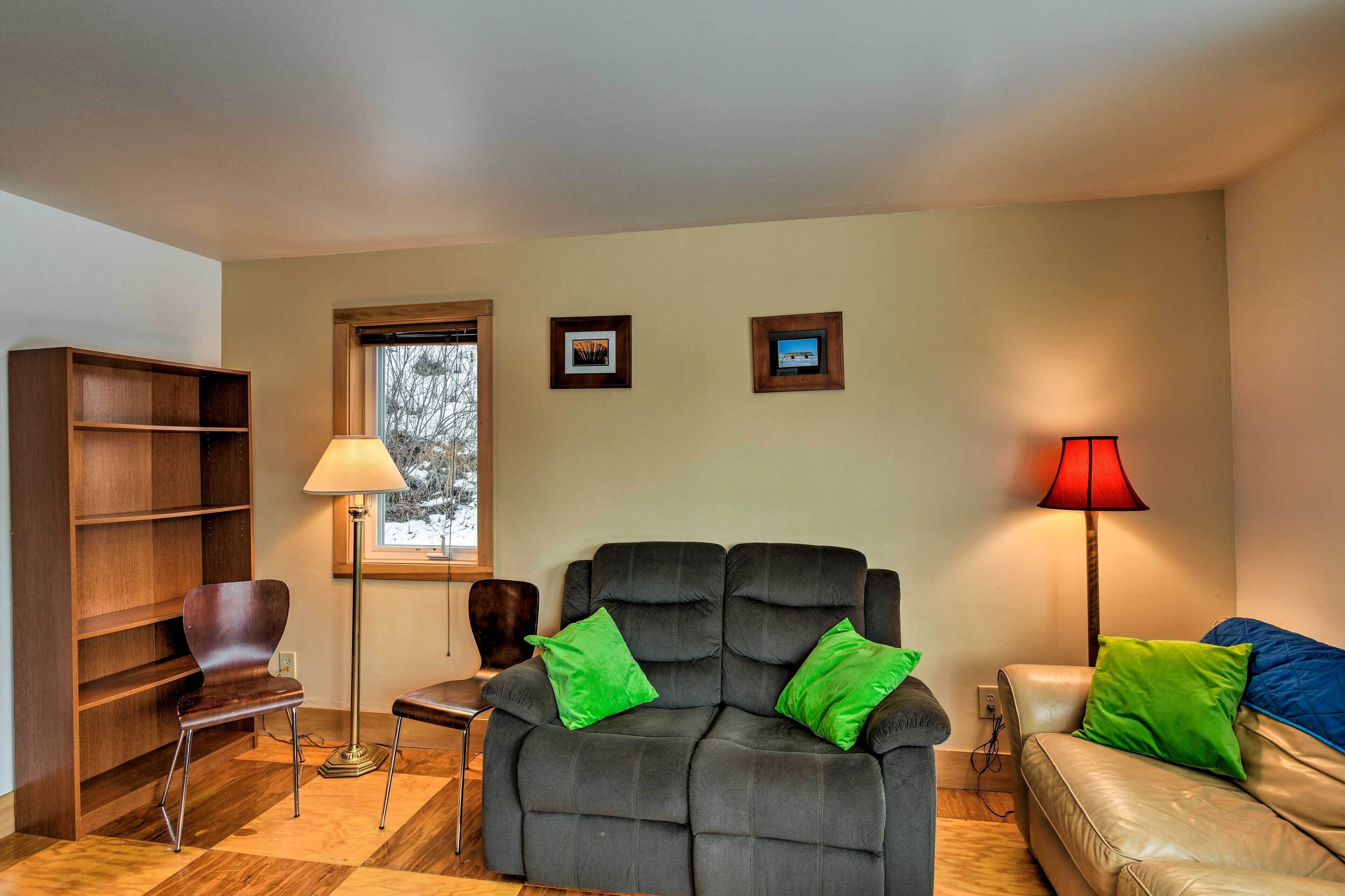 A living room with a dark sofa, two green pillows, a wooden bookshelf, a lamp, and a window.
