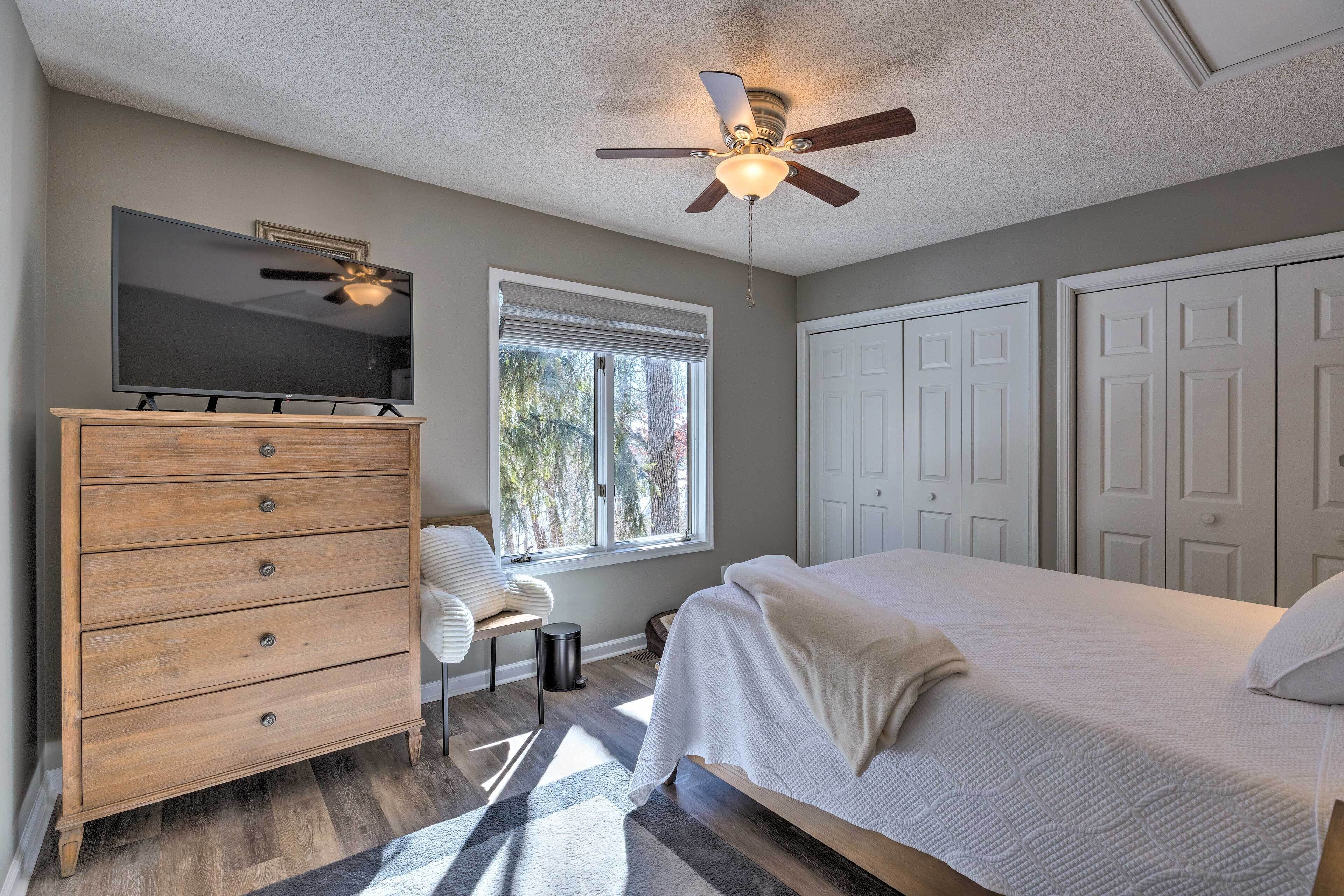 Une chambre à coucher comprenant un lit, une commode, un téléviseur, un ventilateur de plafond et une fenêtre avec des stores.