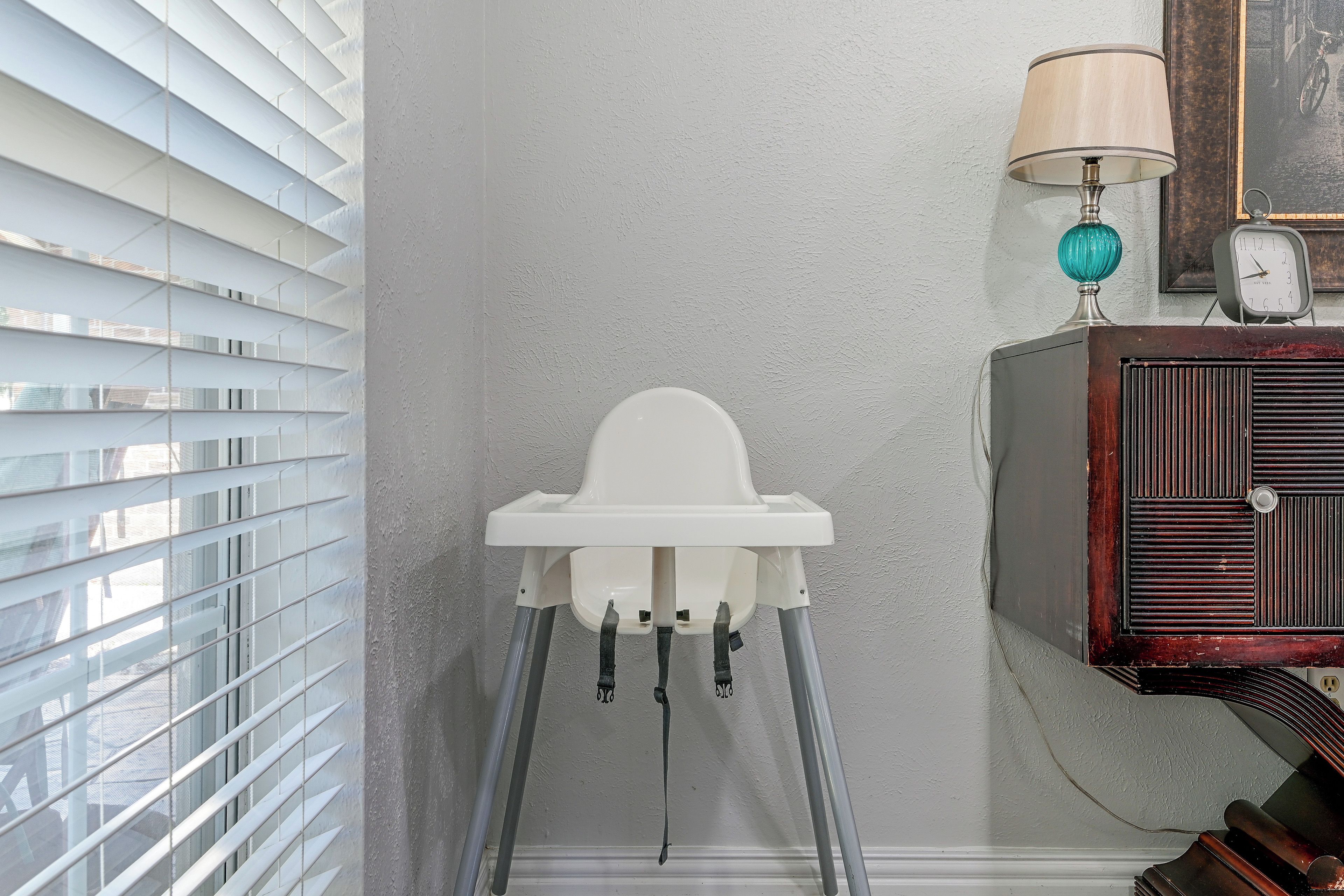 A bathroom with a white pedestal sink, a wall-mounted clock, and a window with blinds.