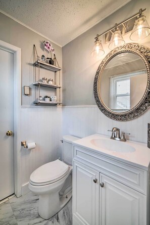 A bathroom with a white toilet, vanity with cabinets, a mirror with an ornate frame, and shelves with decorative items. - Coastal Brigantine Retreat - Walk to Beach! (Brigantine)