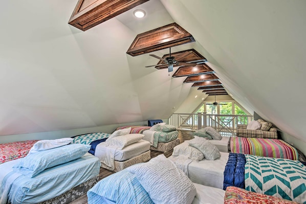 Une chambre confortable située dans les combles, avec plusieurs lits, un ventilateur de plafond et une vue sur l’extérieur par une fenêtre.
