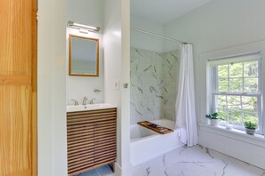 Une salle de bain avec un meuble-lavabo en bois, une baignoire, une douche avec un mur en marbre et une fenêtre agrémentée de plantes.