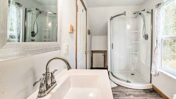 Une salle de bain avec un lavabo blanc, une cabine de douche en verre et une fenêtre laissant entrer la lumière naturelle.