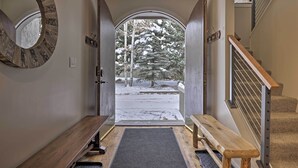 A hallway with a wooden bench, a round mirror, and a view of a snowy landscape through an open door. - Townhome ~ 4 Mi to Beaver Creek & 8 Mi to Vail! (Avon)