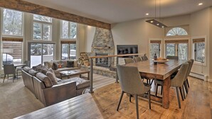 A spacious living area with a large wooden dining table, comfortable seating, a stone fireplace, and large windows offering views of the outdoors. - Townhome ~ 4 Mi to Beaver Creek & 8 Mi to Vail! (Avon)