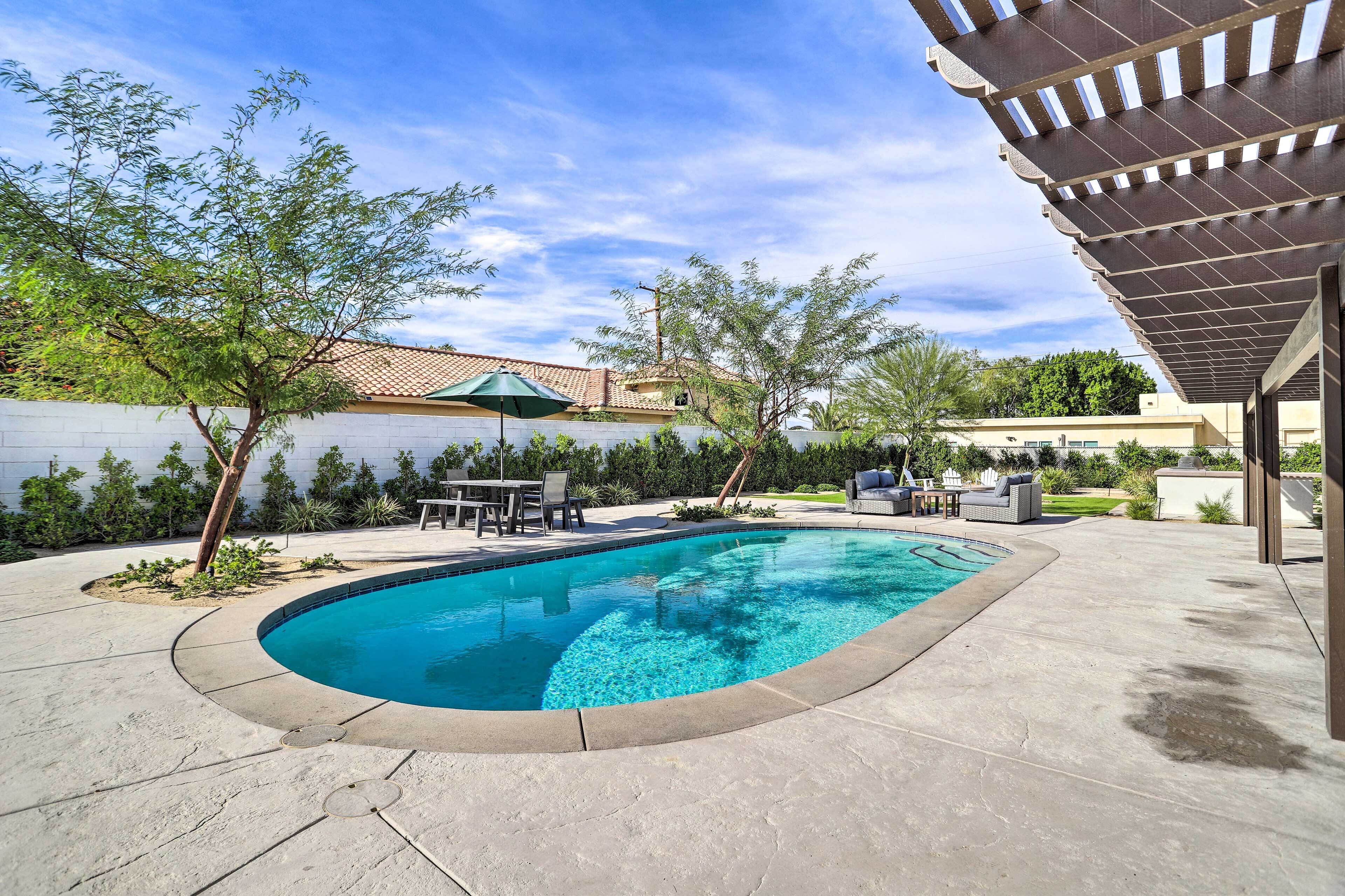 A swimming pool with a curved edge, surrounded by a concrete deck. There are outdoor seating areas and a pergola structure.