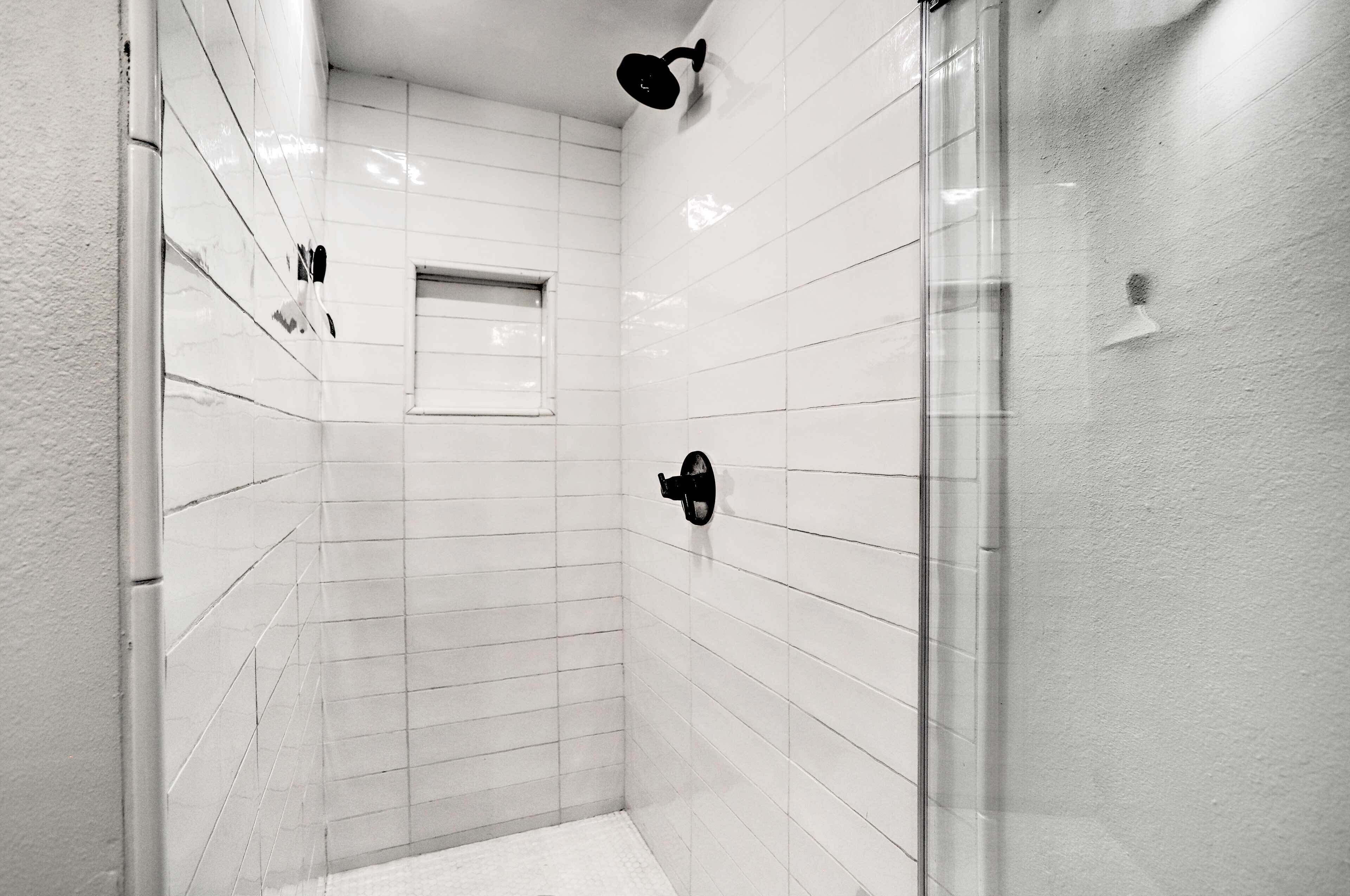 Une salle de bain moderne avec des murs recouverts de carreaux blancs, une douche en verre et une petite fenêtre.