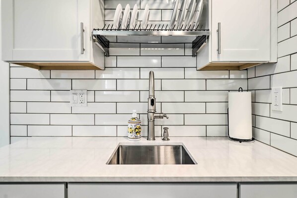 Uma cozinha com painel de azulejos brancos, pia de aço inoxidável e escorredor de louça sobre a bancada.