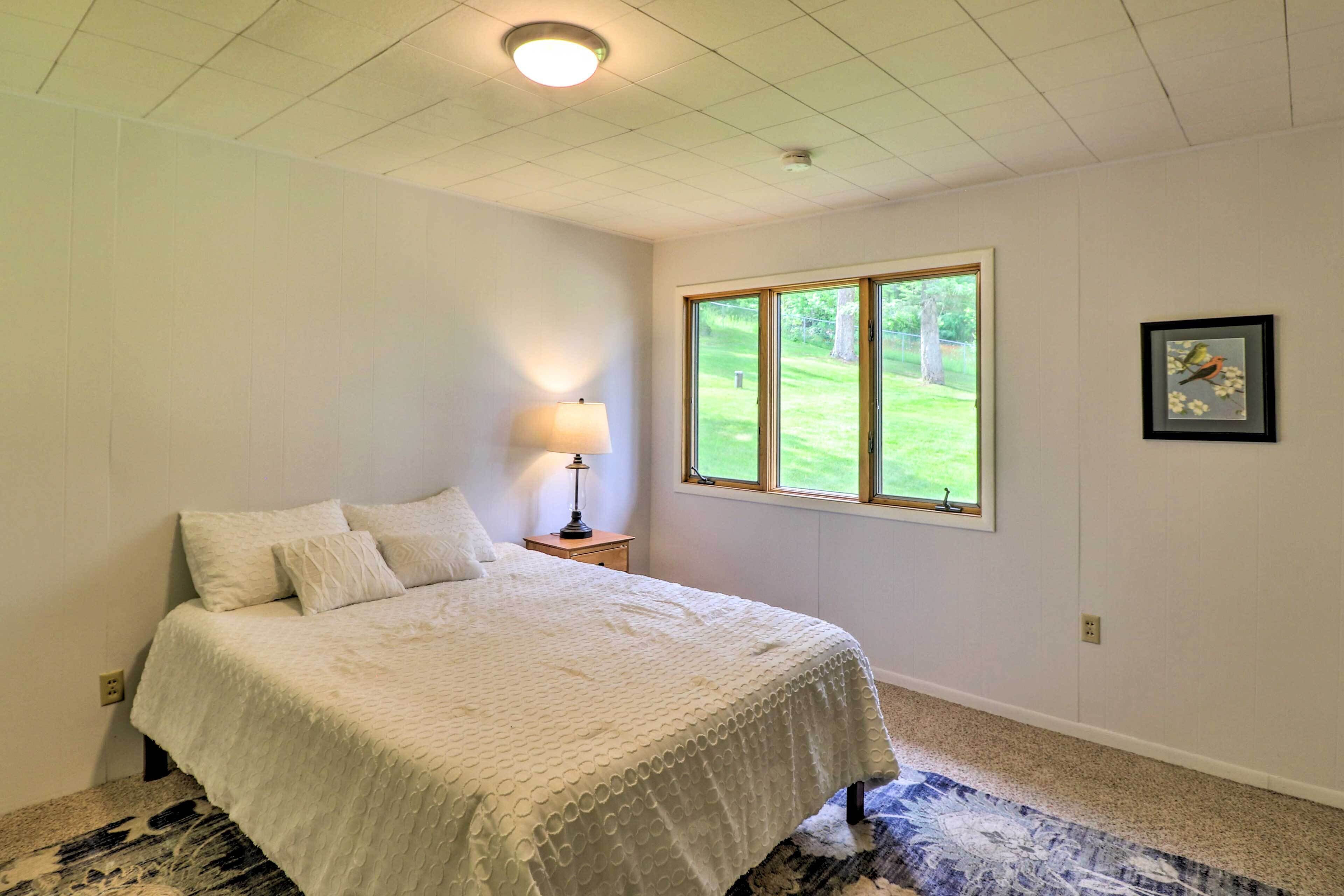 A bedroom with a bed, a nightstand, a lamp, a window with a view of greenery, and a framed picture on the wall.