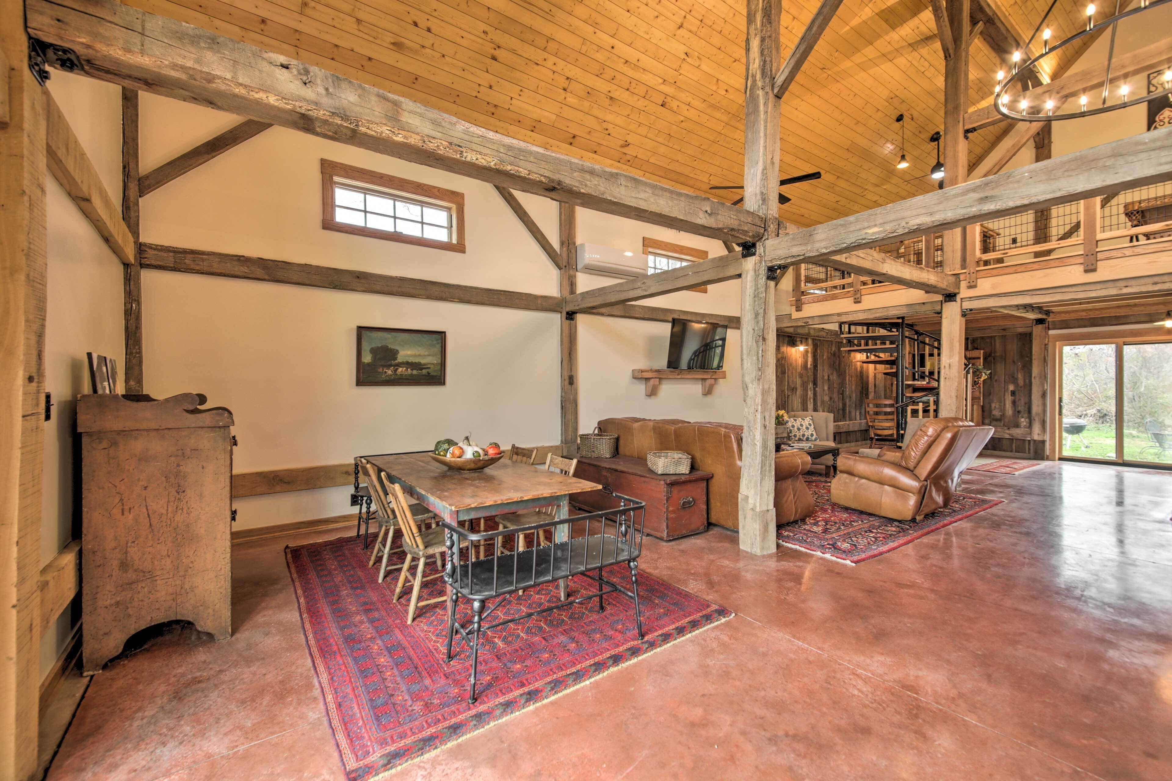 A spacious interior with a high ceiling, wooden beams, a dining area with a table and chairs, a leather sofa, and a large window providing a view of the outdoors.