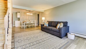 A living room with a grey sofa, patterned rug, and a dining area with white furniture. - Indian Lake Vacation Rental w/ Fire Pit & Sauna (Lakeview)