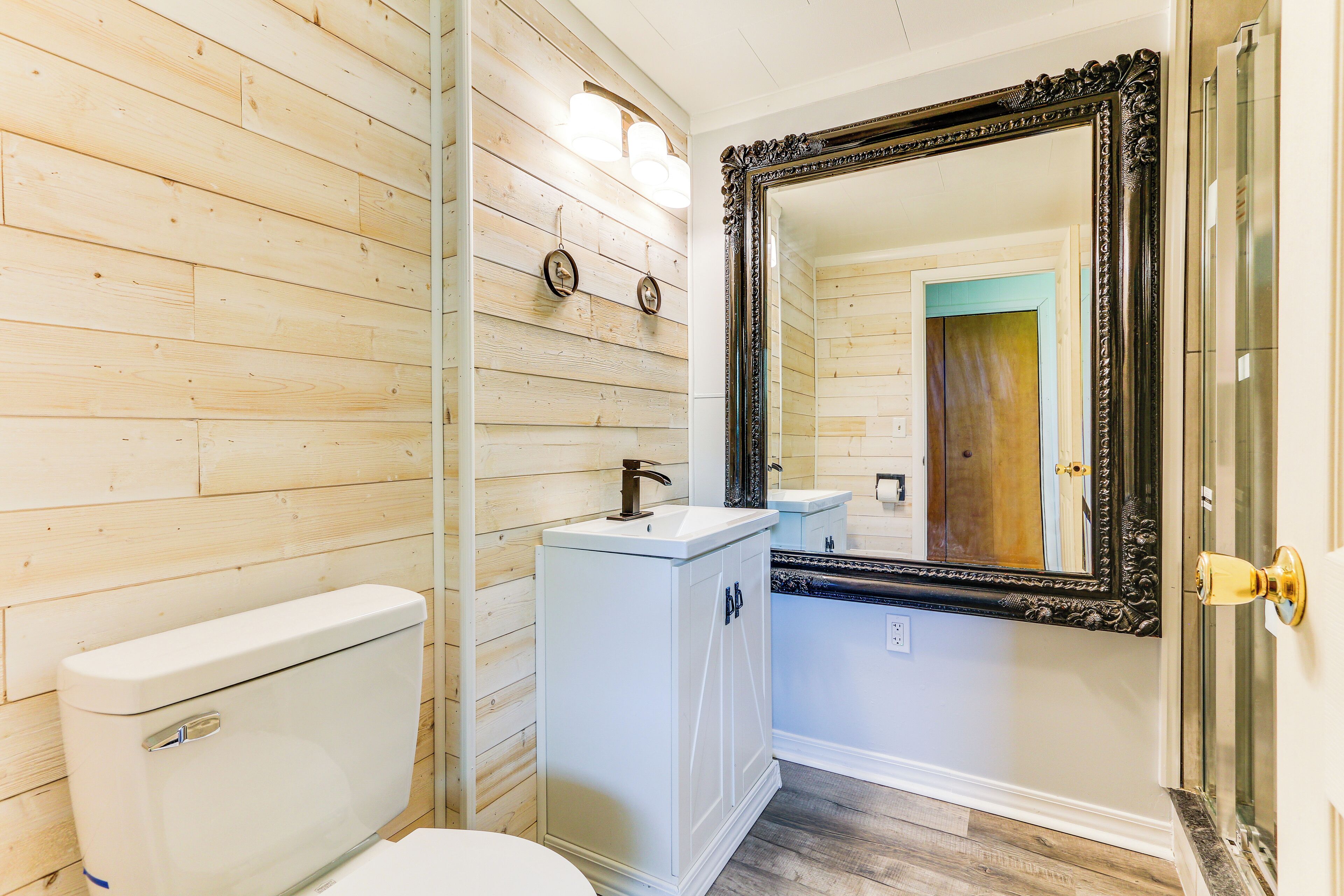 A bathroom with wooden panel walls, a white toilet, a pedestal sink, and a large ornate mirror.
