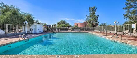 A clear swimming pool with a tiled deck, surrounded by a fence, and a building in the background.