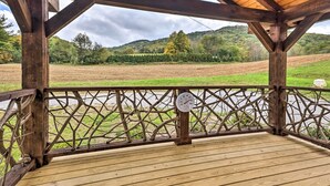 Een houten terras met uitzicht op een rivier en velden, een klok en een metalen hekwerk.