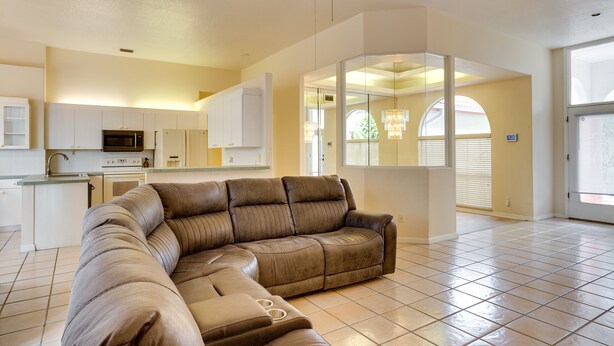 A spacious living area with a large L-shaped sectional sofa, a kitchen with white cabinetry, and a large window providing natural light. - Port St Lucie Home ~ 3 Mi to Botanical Gardens! (Port Saint Lucie)