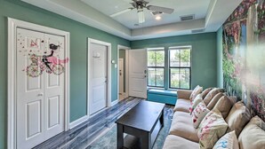 A room with teal walls, a ceiling fan, a window with a view, a sofa with floral cushions, and a door with butterfly decals. - Chic Midtown House ~ 2 Mi to Dtwn Houston! (Houston)