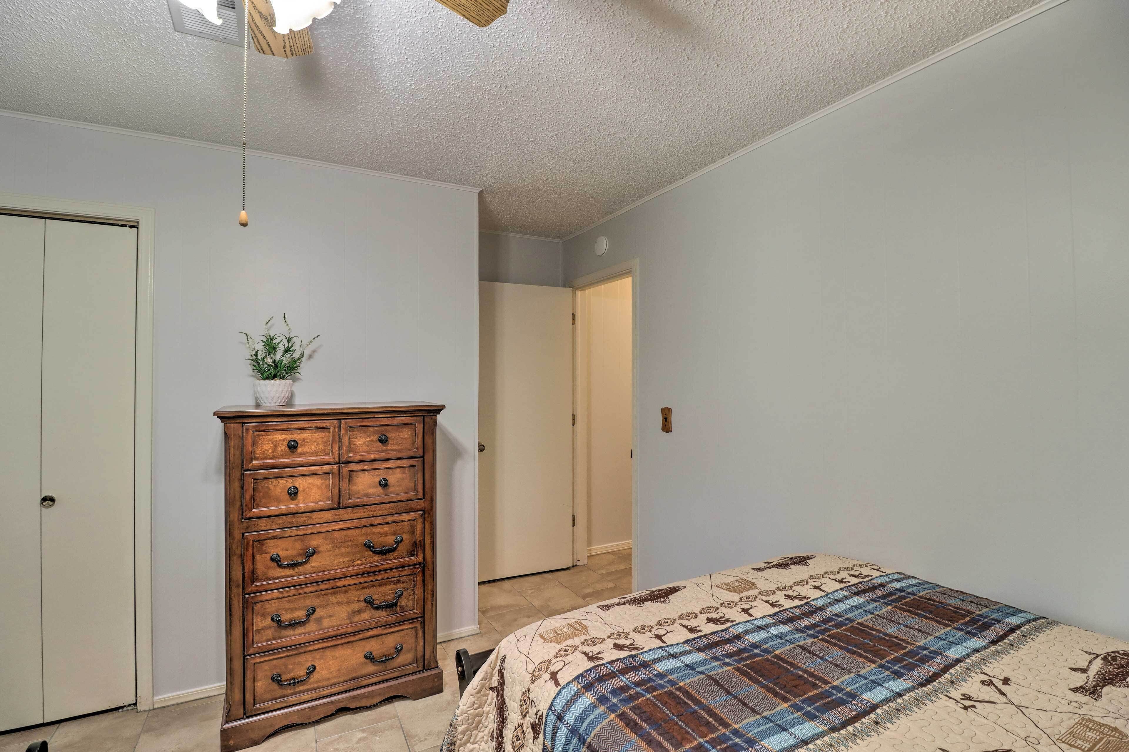 A bedroom with a wooden dresser, a bed with a plaid bedspread, a ceiling fan, and a door leading to another room.