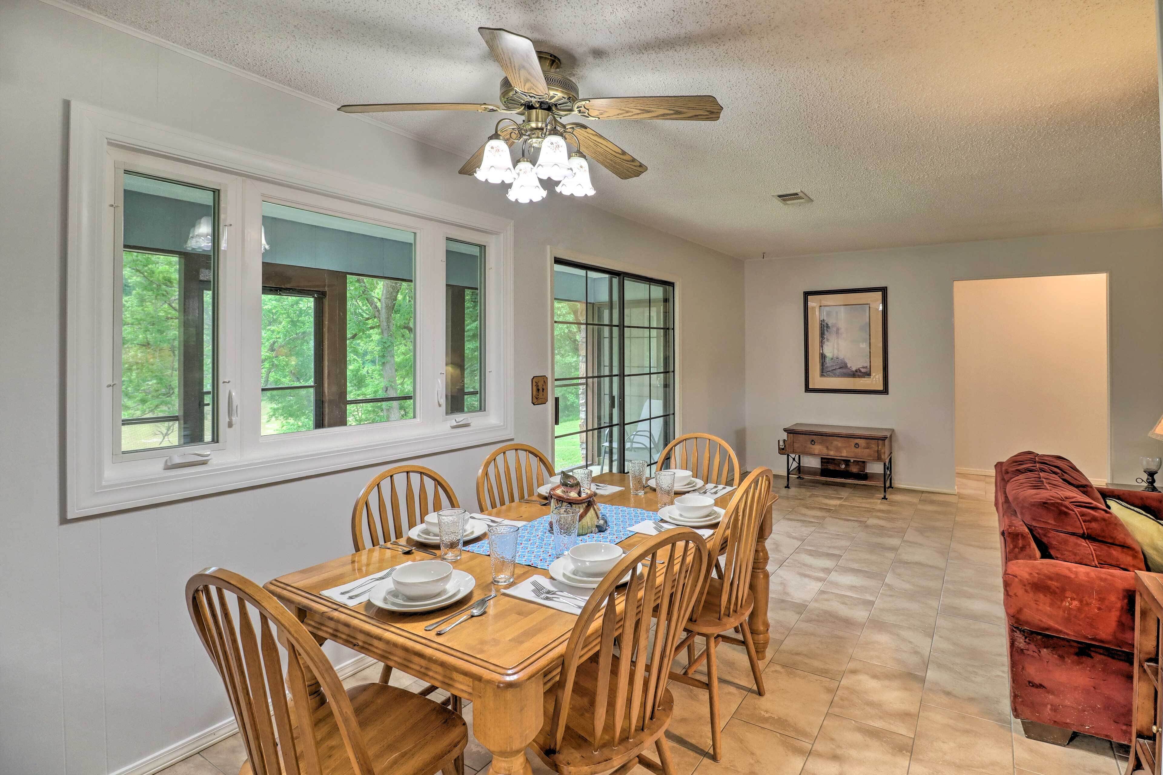 A dining room with a wooden table set for six, large windows, a ceiling fan, and a red sofa in the background.