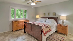 A bedroom with a wooden bed, a ceiling fan, a window with a view of trees, a nightstand with a lamp, and a dresser. - Norfork Getaway: Walk in White River Access! (Norfork)
