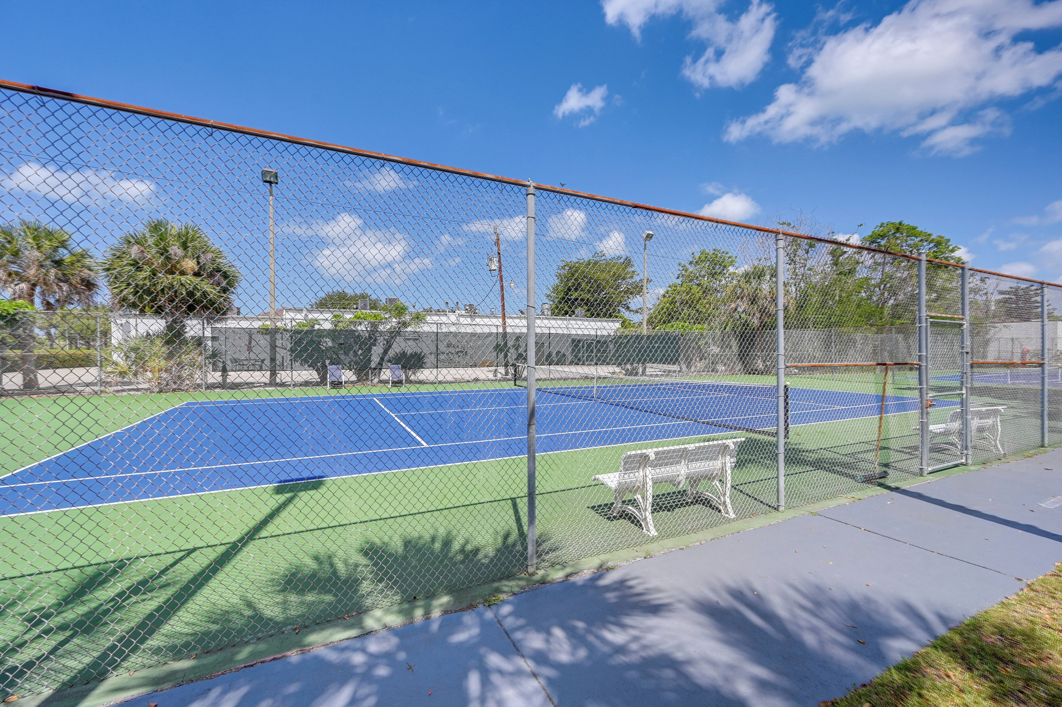 A tennis court with a chain-link fence, benches, and a clear blue sky.