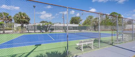 A tennis court with a chain-link fence, benches, and a clear blue sky.