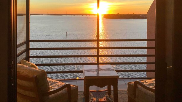 A balcony with a view of the sunset over a body of water, a table, and two chairs.